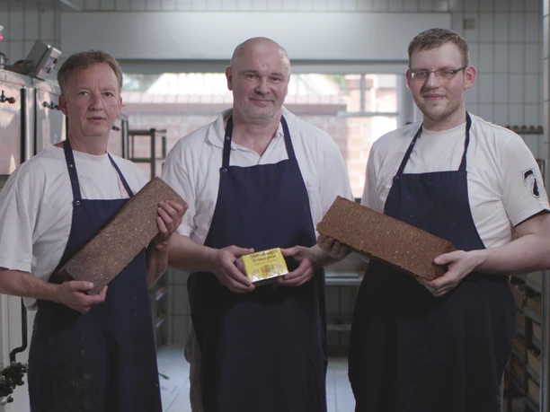 Vollkornbäckerei Ahaus Drei Bäcker in blauen Schürzen stehen in einer Backstube und halten frisch gebackenes Vollkornbrot.