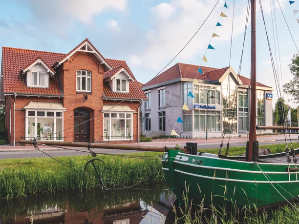 Haus der Kultur, Fehntjer Forum, Rhauderfehn Rotes Backsteingebäude mit einem grünen Plattbodenschiff auf einem Kanal davor bei blauem Himmel.