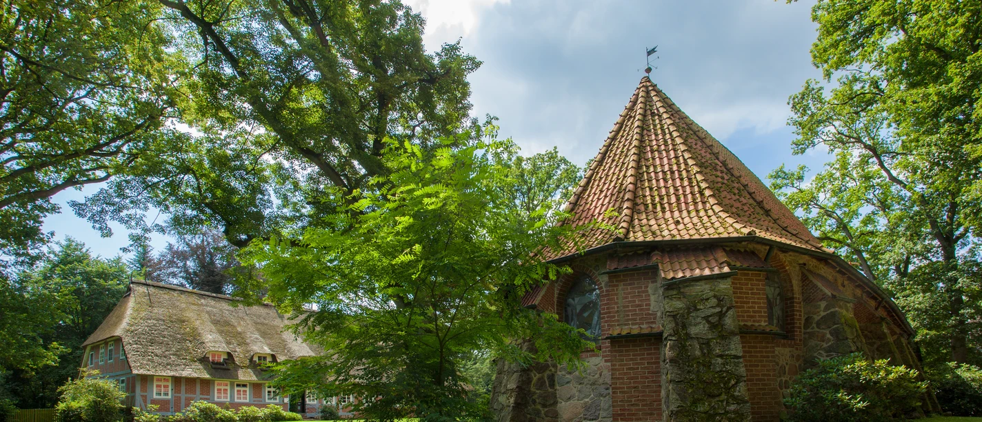 Sommermusik in Bispinger Kirchen Alte Feldsteinkirche mit Ziegeldach und ein Reetdachhaus umgeben von grünen Bäumen.