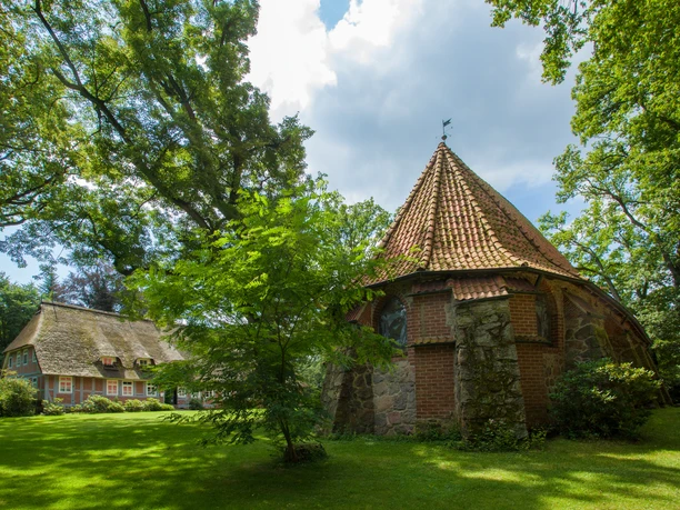 Sommermusik in Bispinger Kirchen Alte Feldsteinkirche mit Ziegeldach und ein Reetdachhaus umgeben von grünen Bäumen.