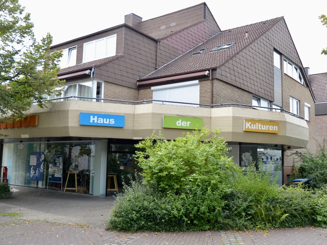 Modernes Gebäude mit Schriftzug "Haus der Kulturen", umgeben von Bäumen und Pflanzen.