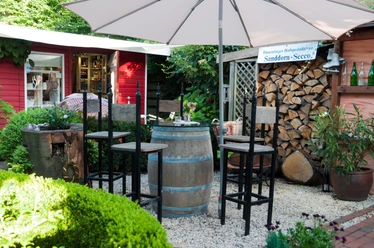 Holztisch aus Weinfass mit hohen Stühlen im Hofgarten, umgeben von Pflanzen und Holzlager.