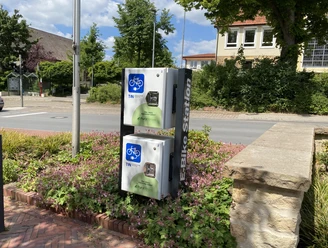 e_bike-ladestation_rathaus_cc_gemeinde-hagen-atw Ladestation für E-Bikes an einer Straßenkreuzung in einer grünen Umgebung.