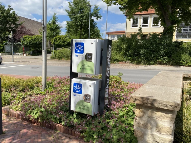 e_bike-ladestation_rathaus_cc_gemeinde-hagen-atw Ladestation für E-Bikes an einer Straßenkreuzung in einer grünen Umgebung.