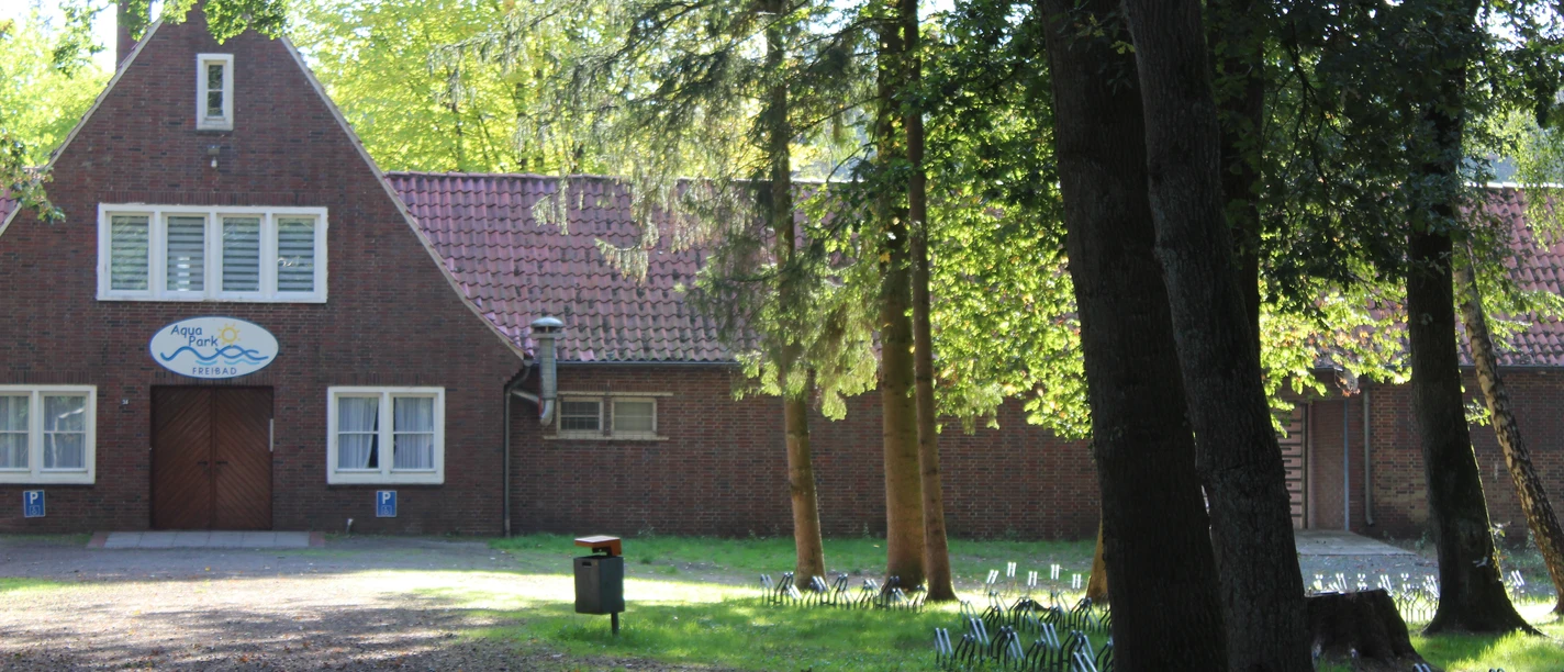 Aqua Park Freibad Papenburg Backsteinbau mit rotem Dach und Aqua-Park-Logo, umgeben von Bäumen im Sonnenlicht.