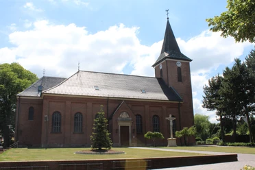 Mariä-Himmelfahrts-Kirche in Lorup Backsteinkirche mit Turm und Kreuz im gepflegten Vorhof, umgeben von Bäumen unter blauem Himmel.