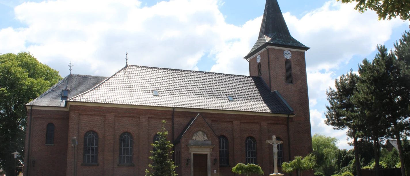Mariä-Himmelfahrts-Kirche in Lorup Backsteinkirche mit Turm und Kreuz im gepflegten Vorhof, umgeben von Bäumen unter blauem Himmel.