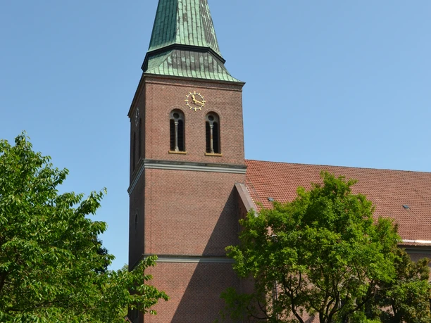 St.-Sixtus-Kirche in Werlte Backsteinkirche mit hohem Turm und grünem Spitzdach, umgeben von Bäumen unter blauem Himmel.