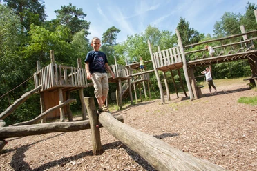 abenteuerspielplatz-bispingen-kinder De avonturenspeeltuin in Bispingen laat lachende kinderen zien die balanceren en klimmen op houten speeltoestellen omringd door bos.