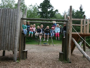 kinder-abenteuerspielplatz Eine fröhliche Gruppe von Kindern sitzt auf der oberen Plattform eines Abenteuerspielplatz-Klettergerüsts aus Holz, vor einem bewaldeten Hintergrund.A cheerful group of children sits on the upper platform of a wooden adventure playground climbing frame, against a wooded background.En munter gruppe børn sidder på den øverste platform af et klatrestativ i træ på en eventyrlig legeplads mod en skovklædt baggrund.Een vrolijke groep kinderen zit op het bovenste platform van een houten klimrek in een avonturenspeeltuin, tegen een beboste achtergrond.