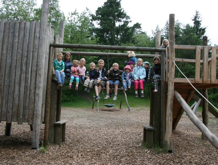 kinder-abenteuerspielplatz Eine fröhliche Gruppe von Kindern sitzt auf der oberen Plattform eines Abenteuerspielplatz-Klettergerüsts aus Holz, vor einem bewaldeten Hintergrund.A cheerful group of children sits on the upper platform of a wooden adventure playground climbing frame, against a wooded background.En munter gruppe børn sidder på den øverste platform af et klatrestativ i træ på en eventyrlig legeplads mod en skovklædt baggrund.Een vrolijke groep kinderen zit op het bovenste platform van een houten klimrek in een avonturenspeeltuin, tegen een beboste achtergrond.