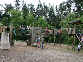abenteuerspielplatz-luhegrund Ein großer Holzspielplatz mit Rutschen, Kletterstrukturen und spielenden Kindern, umgeben von Bäumen.A large wooden playground with slides, climbing structures and children playing, surrounded by trees.En stor trælegeplads med rutsjebaner, klatrestativer og legende børn, omgivet af træer.Een grote houten speeltuin met glijbanen, klimstructuren en spelende kinderen, omringd door bomen.