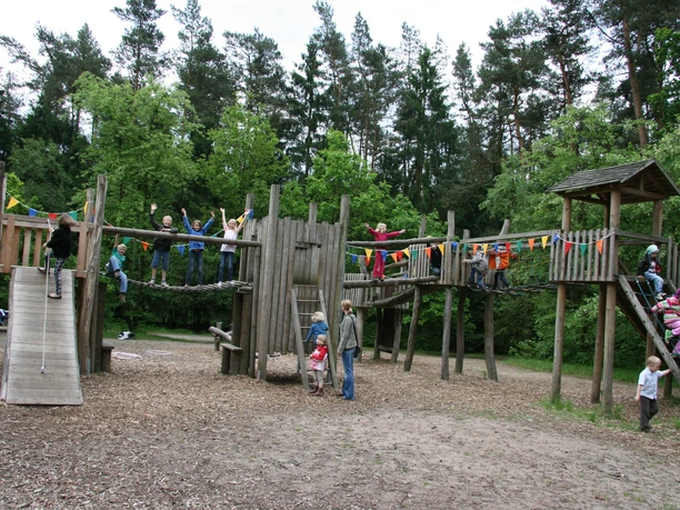 abenteuerspielplatz-luhegrund A large wooden playground with slides, climbing structures and children playing, surrounded by trees.