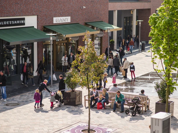 kleiner-überblick Ein belebter Platz mit modernen Geschäften, Menschen flanieren zwischen Boutiquen, Bänken und jungen Bäumen.