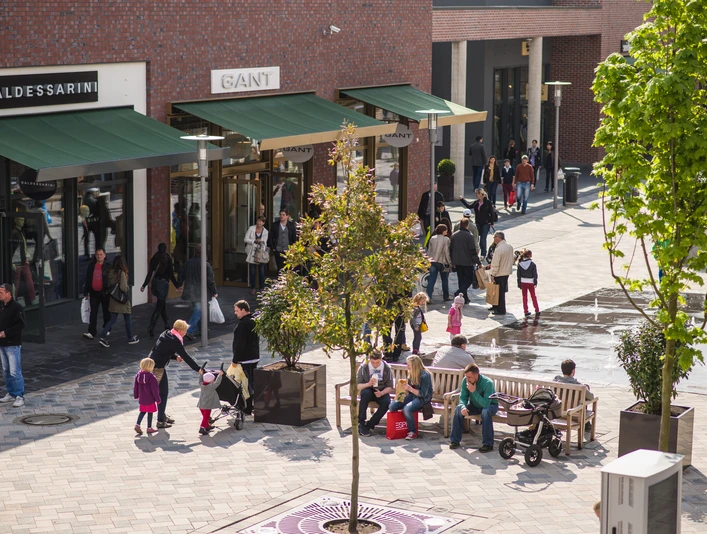 kleiner-überblick Ein belebter Platz mit modernen Geschäften, Menschen flanieren zwischen Boutiquen, Bänken und jungen Bäumen.