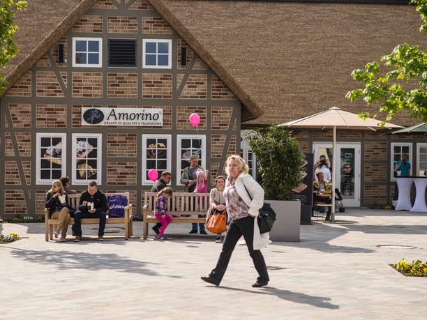 designer-outlet-soltau Im Designer Outlet Soltau sitzen Menschen auf Bänken vor einem Fachwerkgebäude mit einem Café und genießen das sonnige Wetter.