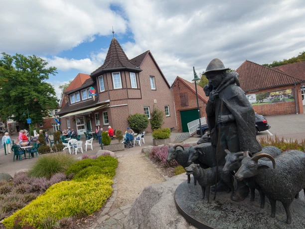 bispingen-ortsmitte-eisdiele De foto van het centrum van Bispingen toont een idyllisch tafereel: Een ijssalon met kleurrijke parasols voor een bakstenen gebouw. Rechts op de voorgrond staat een bronzen beeldhouwwerk van een historische figuur met schapen. Mensen genieten van het zachte weer en kletsen aan de buitentafels. Dit geeft het dorpscentrum een uitnodigende en levendige sfeer.