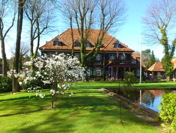 bispingen-iserhatsche Ein historisches, rotes Backsteinhaus mit Satteldach, umgeben von blühenden Bäumen und einem Teich.