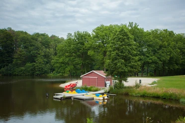 am-brunausee Ein idyllischer See mit einem roten Boots- und Lagerhaus, umgeben von grünen Bäumen und einem Steg mit bunten Tretbooten, vermittelt Ruhe und Naturschönheit.