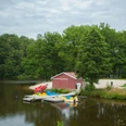 am-brunausee Ein idyllischer See mit einem roten Boots- und Lagerhaus, umgeben von grünen Bäumen und einem Steg mit bunten Tretbooten, vermittelt Ruhe und Naturschönheit.