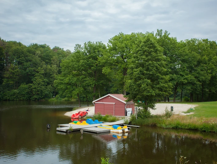 am-brunausee Ein idyllischer See mit einem roten Boots- und Lagerhaus, umgeben von grünen Bäumen und einem Steg mit bunten Tretbooten, vermittelt Ruhe und Naturschönheit.