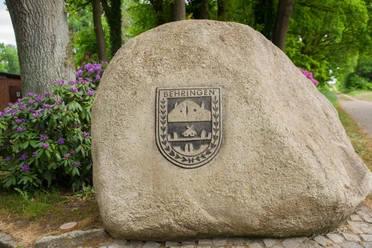 Schützenfest in Behringen Ein großer Findling mit dem Wappen von Behringen, umgeben von Bäumen und Rhododendren.