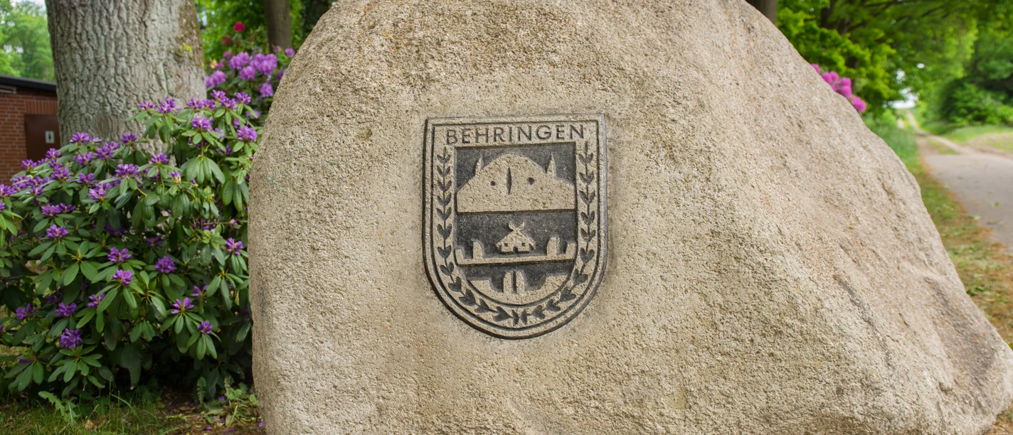Schützenfest in Behringen Ein großer Findling mit dem Wappen von Behringen, umgeben von Bäumen und Rhododendren.