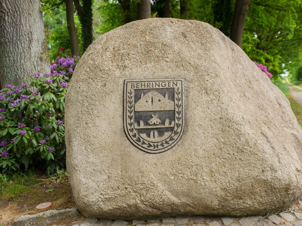 Schützenfest in Behringen Ein großer Findling mit dem Wappen von Behringen, umgeben von Bäumen und Rhododendren.