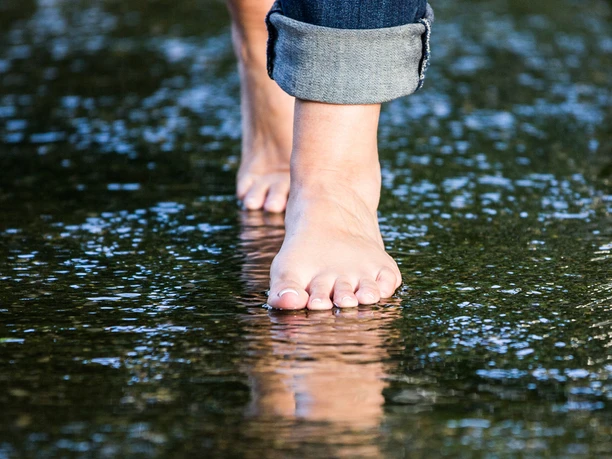 barfusspark-fuesse Barfüßige Person schreitet durch seichtes Wasser, umgeben von reflektierendem Licht auf der Oberfläche.