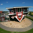 Das verrückte Haus Aussenansicht Das Bild zeigt ein auf dem Kopf stehendes, rot-weißes Holzhaus, umgeben von grünem Rasen und einer asphaltierten Straße.The picture shows a red and white wooden house standing on its head, surrounded by a green lawn and an asphalt road.Billedet viser et rødt og hvidt træhus, der står på hovedet, omgivet af en grøn græsplæne og en asfalteret vej.De foto toont een rood-wit houten huis dat op zijn kop staat, omringd door een groen grasveld en een geasfalteerde weg.