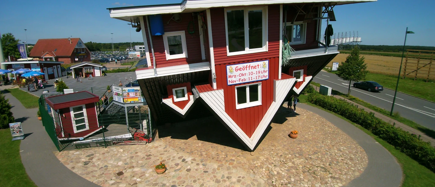 Das verrückte Haus Aussenansicht The picture shows a red and white wooden house standing on its head, surrounded by a green lawn and an asphalt road.