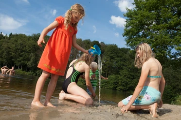 brunausee-schwimmen Eine Gruppe von Kindern spielt fröhlich am Strand, während ein Mädchen mit rotem Kleid Wasser gießt.