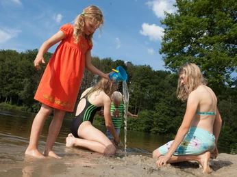 brunausee-schwimmen Eine Gruppe von Kindern spielt fröhlich am Strand, während ein Mädchen mit rotem Kleid Wasser gießt.