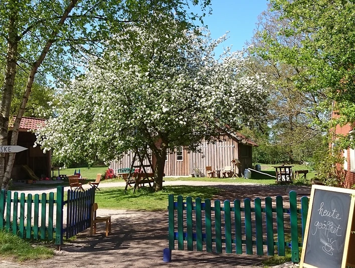 Melkhuske Schatteburg, Gemeinde Rhauderfehn, Ostfriesland Grüner Holzzaun öffnet sich zu einem Garten mit blühendem Baum, Holztischen und einem Schild mit der Aufschrift - Heute geöffnet