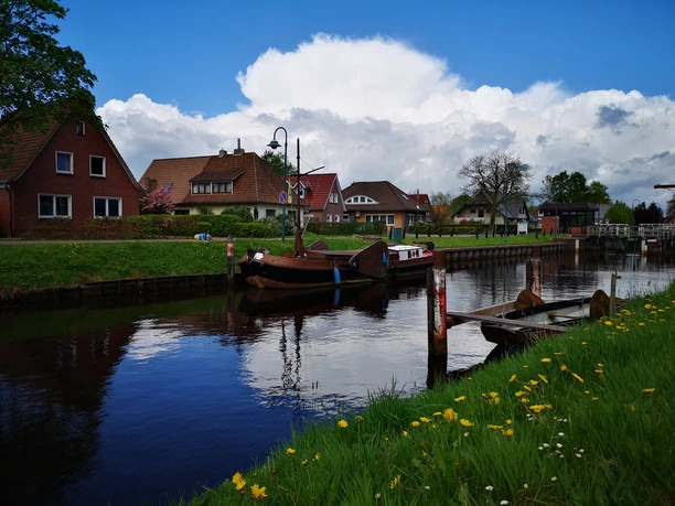 Torfschiff Hoffnung - Rhauderfehn - Ostfriesland 2 Kleines Dorf mit Häusern am Ufer eines ruhigen Kanals, Segelboot im Wasser, grünes Gras mit gelben Blumen im Vordergrund.