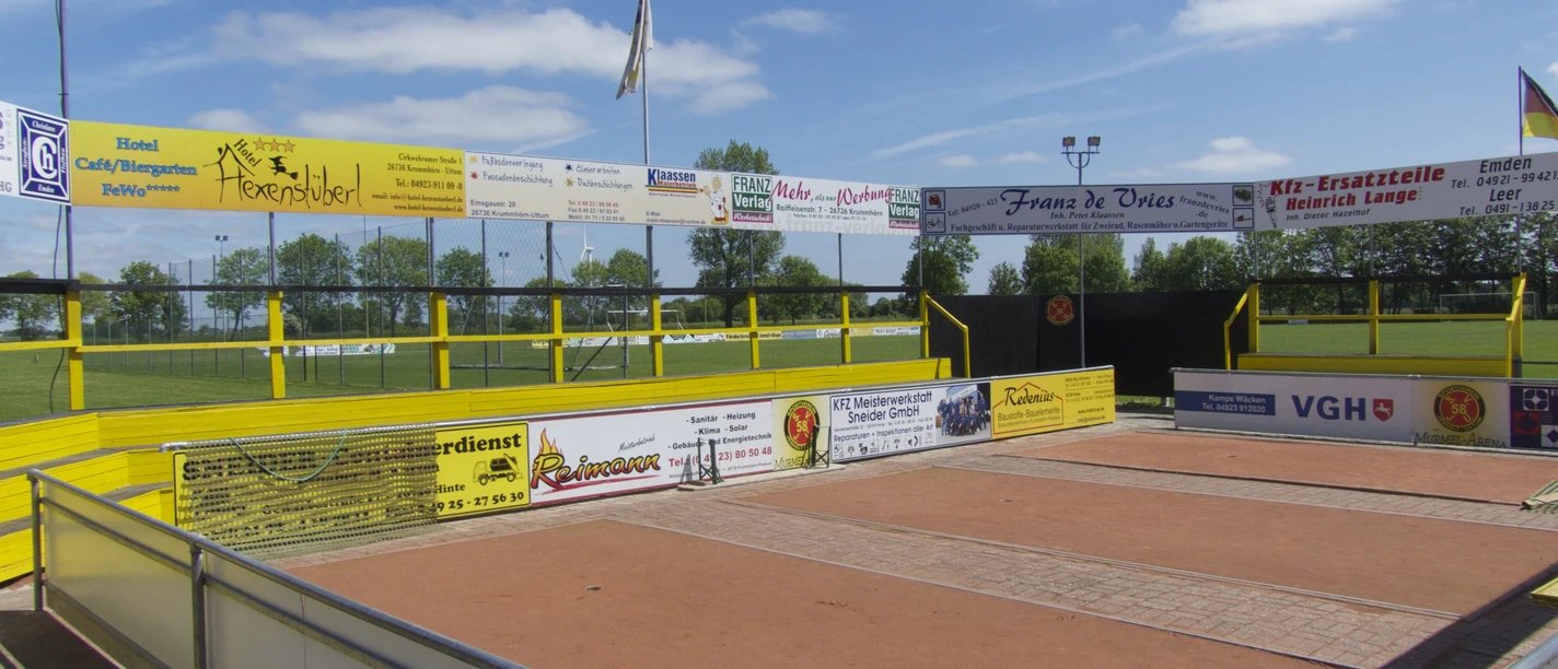 Murmelarena in Uttum Sandige Spielfläche der Murmelarena mit Werbebanden neben einem grünen Sportplatz.