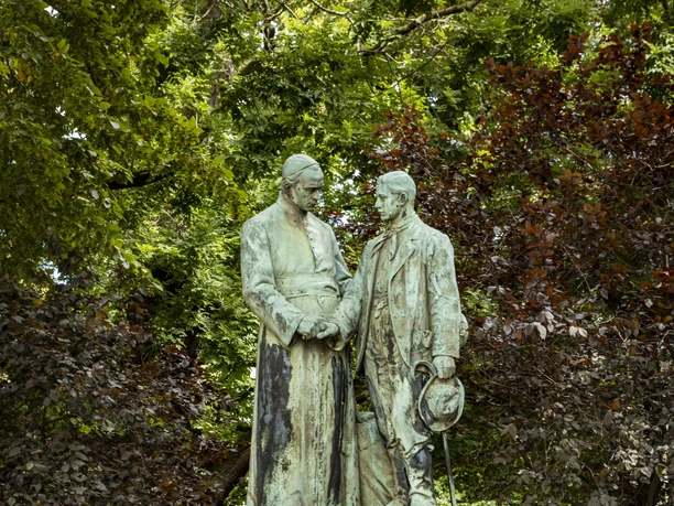 Adolf Kolping Monument The monument shows Adolf Kolping and a young man in typical 19th century working-class clothing. Both are standing in the stylish park, surrounded by dense foliage and a flower border. The monument is mounted on a pedestal on which the name "ADOLF KOLPING" is engraved.