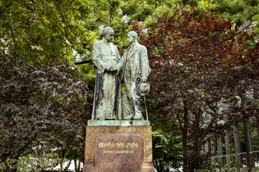 Adolf Kolping Denkmal Adolf Kolping Denkmal, bestehend aus zwei respektvoll gestalteten Bronzestatuen, umgeben von üppigen Bäumen.