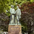 Adolf Kolping Monument Adolf Kolping Denkmal, bestehend aus zwei respektvoll gestalteten Bronzestatuen, umgeben von üppigen Bäumen.Adolf Kolping monument, consisting of two respectfully designed bronze statues, surrounded by lush trees.