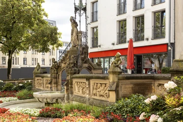 Heinzelmaennchenbrunnen Eine üppige Blumengestaltung umgibt den Heinzelmännchenbrunnen mit seinen detailreichen Reliefs und Skulpturen, dahinter moderne Gebäude.