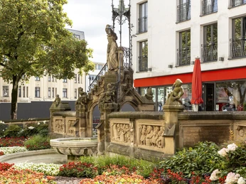 Heinzelmaennchenbrunnen Eine üppige Blumengestaltung umgibt den Heinzelmännchenbrunnen mit seinen detailreichen Reliefs und Skulpturen, dahinter moderne Gebäude.A lush floral design surrounds the Heinzelmännchenbrunnen fountain with its detailed reliefs and sculptures, with modern buildings behind it.
