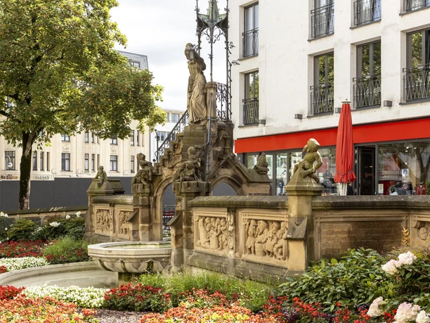 Heinzelmaennchenbrunnen Eine üppige Blumengestaltung umgibt den Heinzelmännchenbrunnen mit seinen detailreichen Reliefs und Skulpturen, dahinter moderne Gebäude.