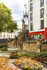 Pixies’ Fountain Heinzelmännchenbrunnen in Cologne: fountain with figures and reliefs, surrounded by flowers.
