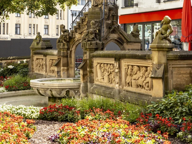 Pixies’ Fountain Heinzelmännchenbrunnen in Cologne: fountain with figures and reliefs, surrounded by flowers.