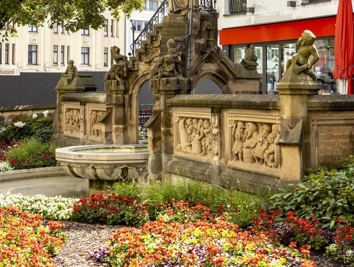 Pixies’ Fountain Heinzelmännchenbrunnen in Köln: Brunnen mit Figuren und Reliefs, umgeben von Blumen.Heinzelmännchenbrunnen in Cologne: fountain with figures and reliefs, surrounded by flowers.