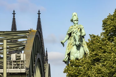 Reiterstandbild König Friedrich Wilhelm IV. Das Reiterstandbild König Friedrich Wilhelm IV. zeigt eine majestätische Bronzestatue auf einem Pferd. Im Hintergrund erhebt sich das Kölner Wahrzeichen, der Dom, während das Bild von der ikonischen Hohenzollernbrücke eingerahmt wird.