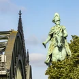 Reiterstandbild König Friedrich Wilhelm IV. Das Reiterstandbild König Friedrich Wilhelm IV. zeigt eine majestätische Bronzestatue auf einem Pferd. Im Hintergrund erhebt sich das Kölner Wahrzeichen, der Dom, während das Bild von der ikonischen Hohenzollernbrücke eingerahmt wird.The equestrian statue of King Frederick William IV shows a majestic bronze statue on a horse. Cologne's landmark, the cathedral, rises in the background, while the image is framed by the iconic Hohenzollern Bridge.