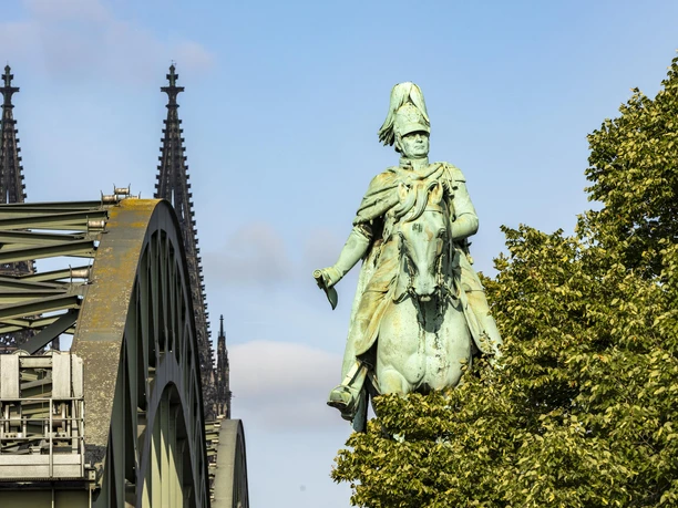 Reiterstandbild König Friedrich Wilhelm IV. Das Reiterstandbild König Friedrich Wilhelm IV. zeigt eine majestätische Bronzestatue auf einem Pferd. Im Hintergrund erhebt sich das Kölner Wahrzeichen, der Dom, während das Bild von der ikonischen Hohenzollernbrücke eingerahmt wird.