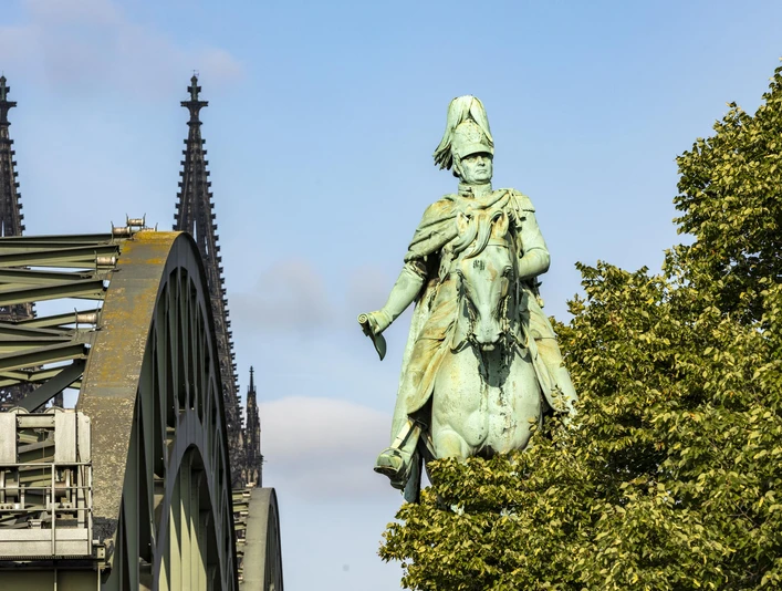 Reiterstandbild König Friedrich Wilhelm IV. Das Reiterstandbild König Friedrich Wilhelm IV. zeigt eine majestätische Bronzestatue auf einem Pferd. Im Hintergrund erhebt sich das Kölner Wahrzeichen, der Dom, während das Bild von der ikonischen Hohenzollernbrücke eingerahmt wird.The equestrian statue of King Frederick William IV shows a majestic bronze statue on a horse. Cologne's landmark, the cathedral, rises in the background, while the image is framed by the iconic Hohenzollern Bridge.