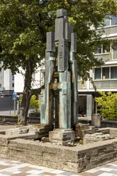 Theo-Burauen-Brunnen Das Bild zeigt den Theo-Burauen-Brunnen, einen eleganten, vertikalen Steingussbrunnen in Köln. Umliegend sind ein grüner Baum und moderne Gebäude sichtbar, die das urbane Stadtbild ergänzen.